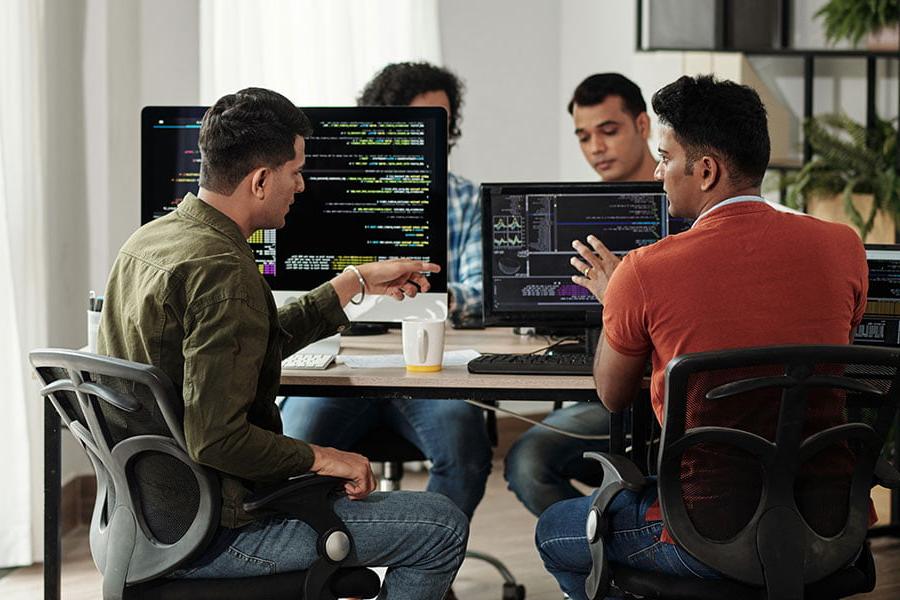 Four individuals sit in front of computers, discussing and pointing at text on the screen