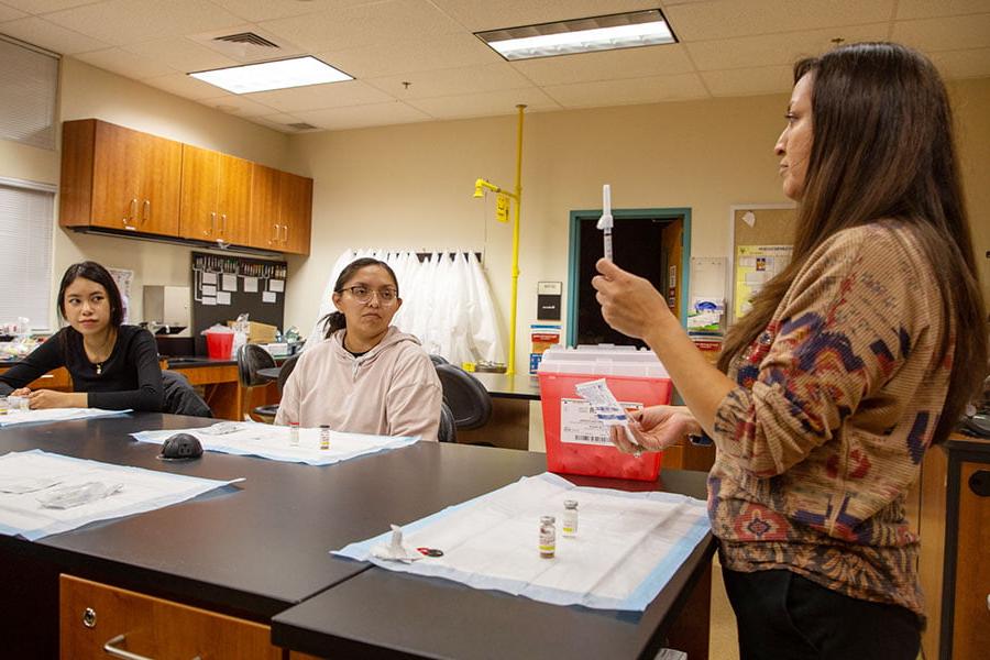 An instructor holds a medical syringe while standing in front of a table while two students look on